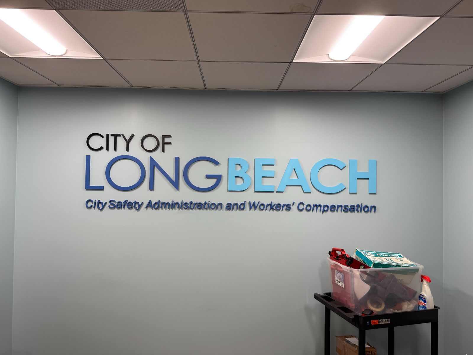 Interior wall with 'City of Long Beach' in dimensional letters: black, blue and light-blue faces above a line reading 'City Safety Administration and Workers' Compensation,' flush-mounted on a pale wall — installed by Hi Sign, Brea, CA