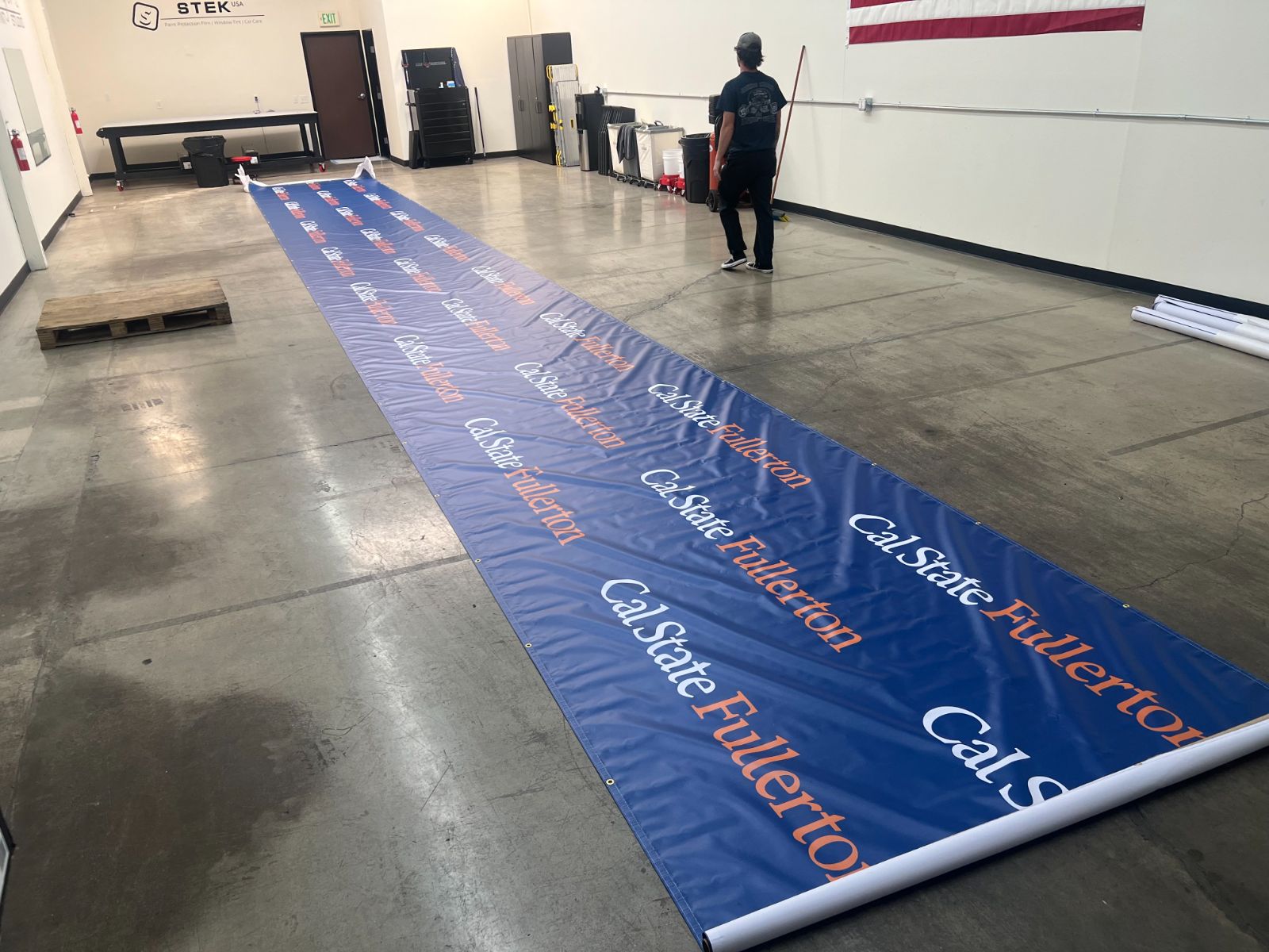 A long Cal State Fullerton step-and-repeat banner unrolled across the shop floor: blue printed vinyl, grommeted edges