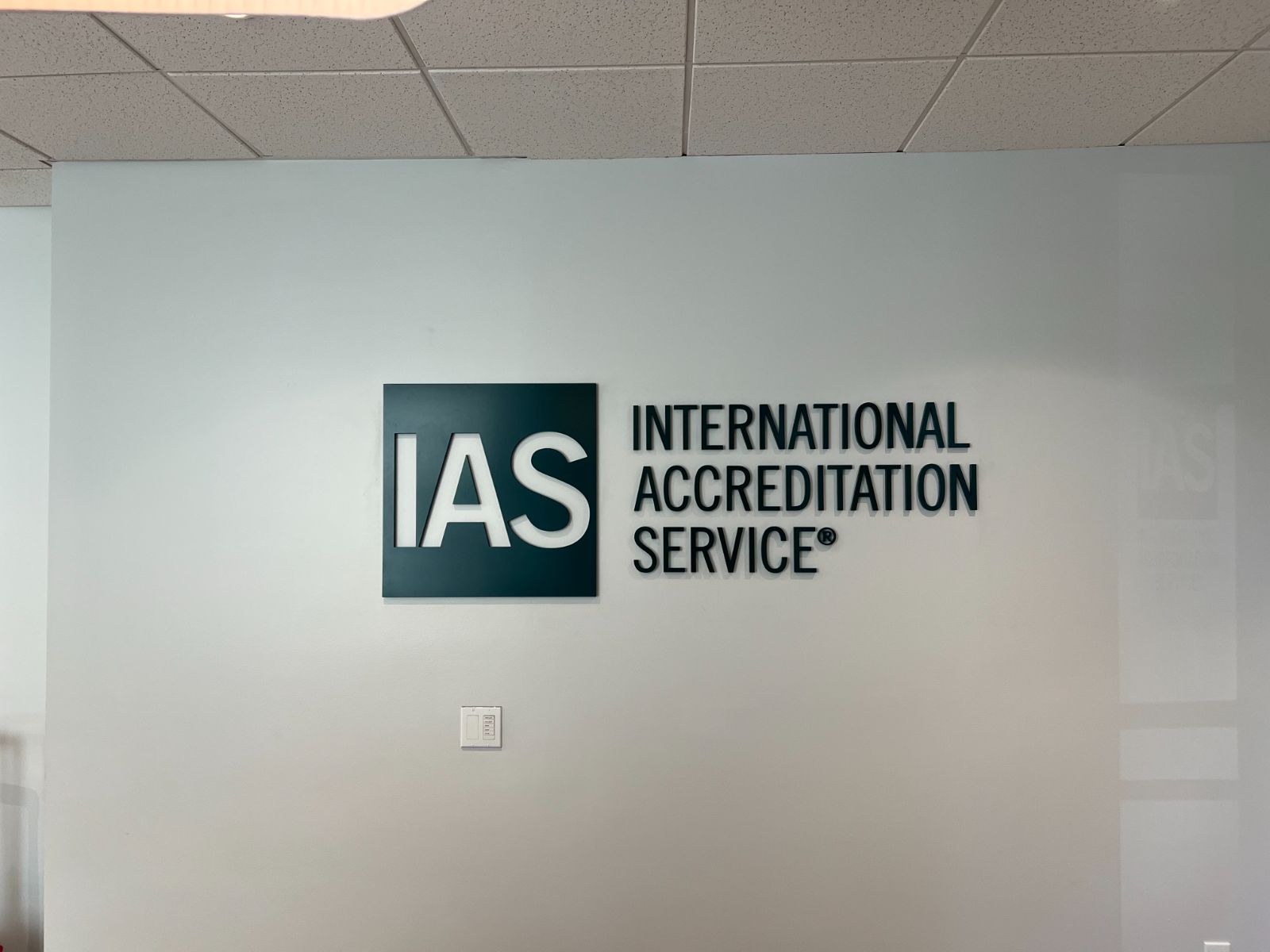 Dimensional lobby logo installed by Hi Sign: IAS (International Accreditation Service) dark teal ACM backer with raised white cast acrylic letterforms, flanked by near-black painted letters, standoff-mounted on a white interior office wall
