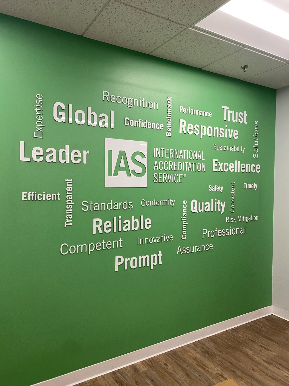 Green office wall covered in a cut-vinyl word cloud of white and grey terms — Global, Leader, Trust, Responsive, Excellence, Reliable, Prompt — around the IAS logo