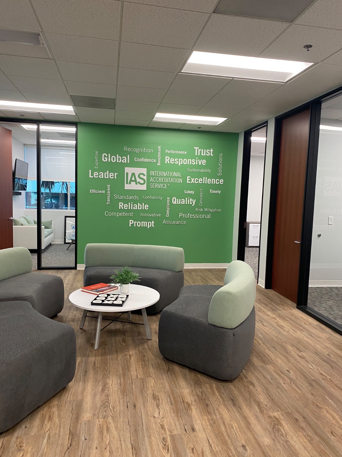 IAS (International Accreditation Service) lobby feature wall: dimensional white IAS logo panel standoff-mounted at the center of a green word-cloud wall graphic — installed by Hi Sign, Brea CA