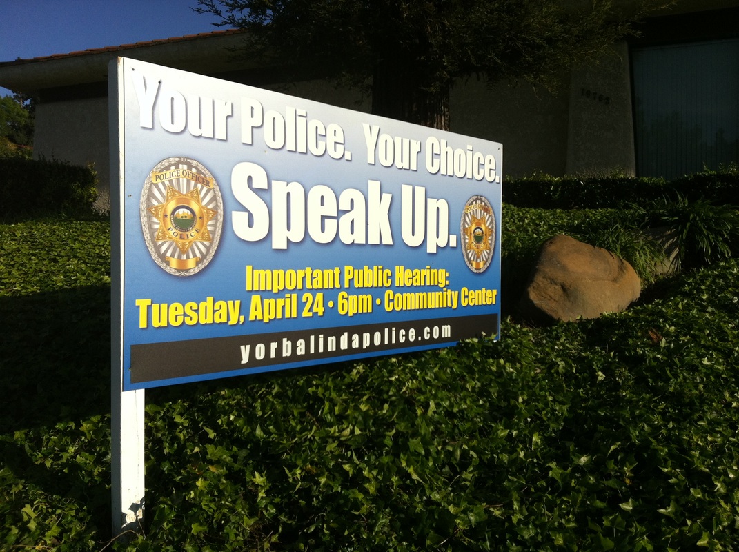 Post-mounted printed panel announcing a Yorba Linda Police public hearing, staked on a landscaped slope